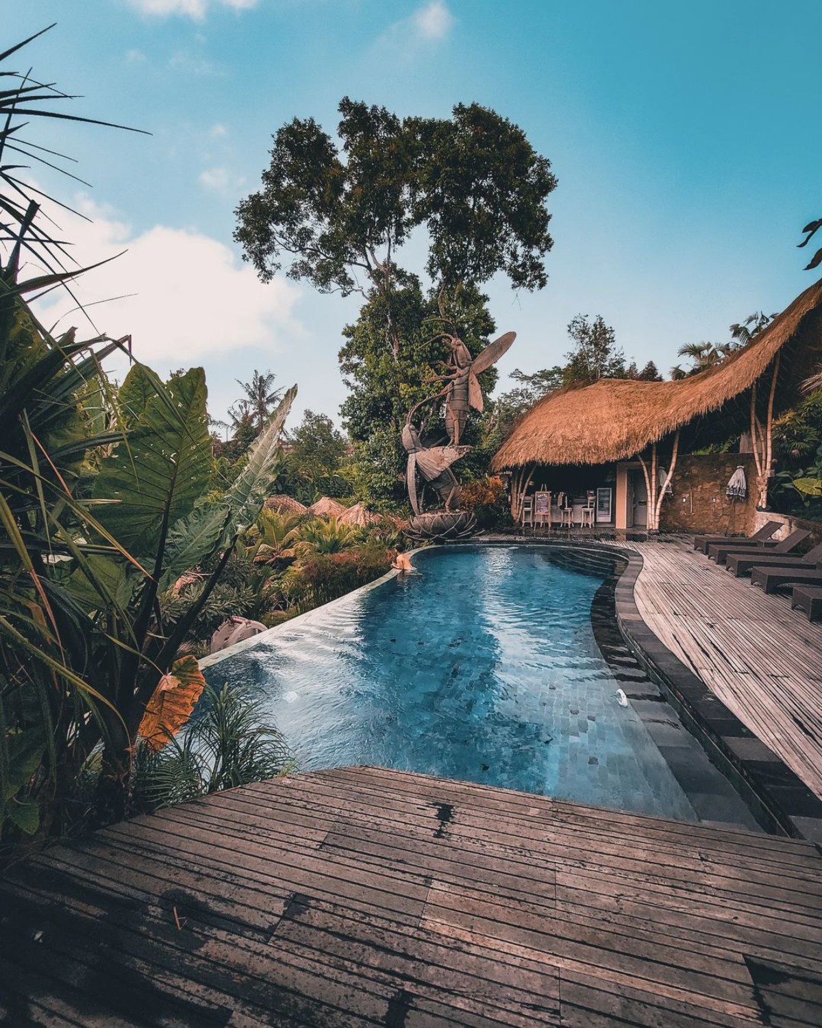 A pristine pool set against a backdrop of thatched huts and tall sculptures, surrounded by lush tropical foliage under a sunlit sky.