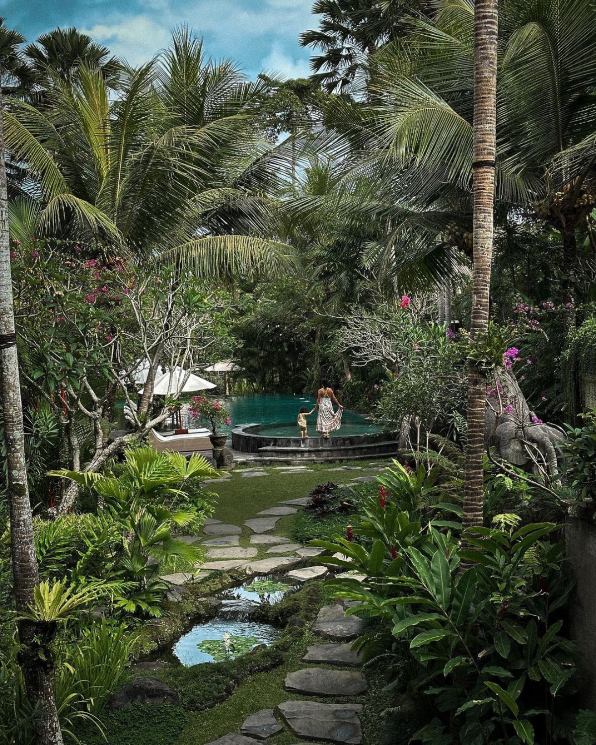 A serene pool surrounded by lush greenery, stone pathways, and a couple enjoying the tranquil setting.