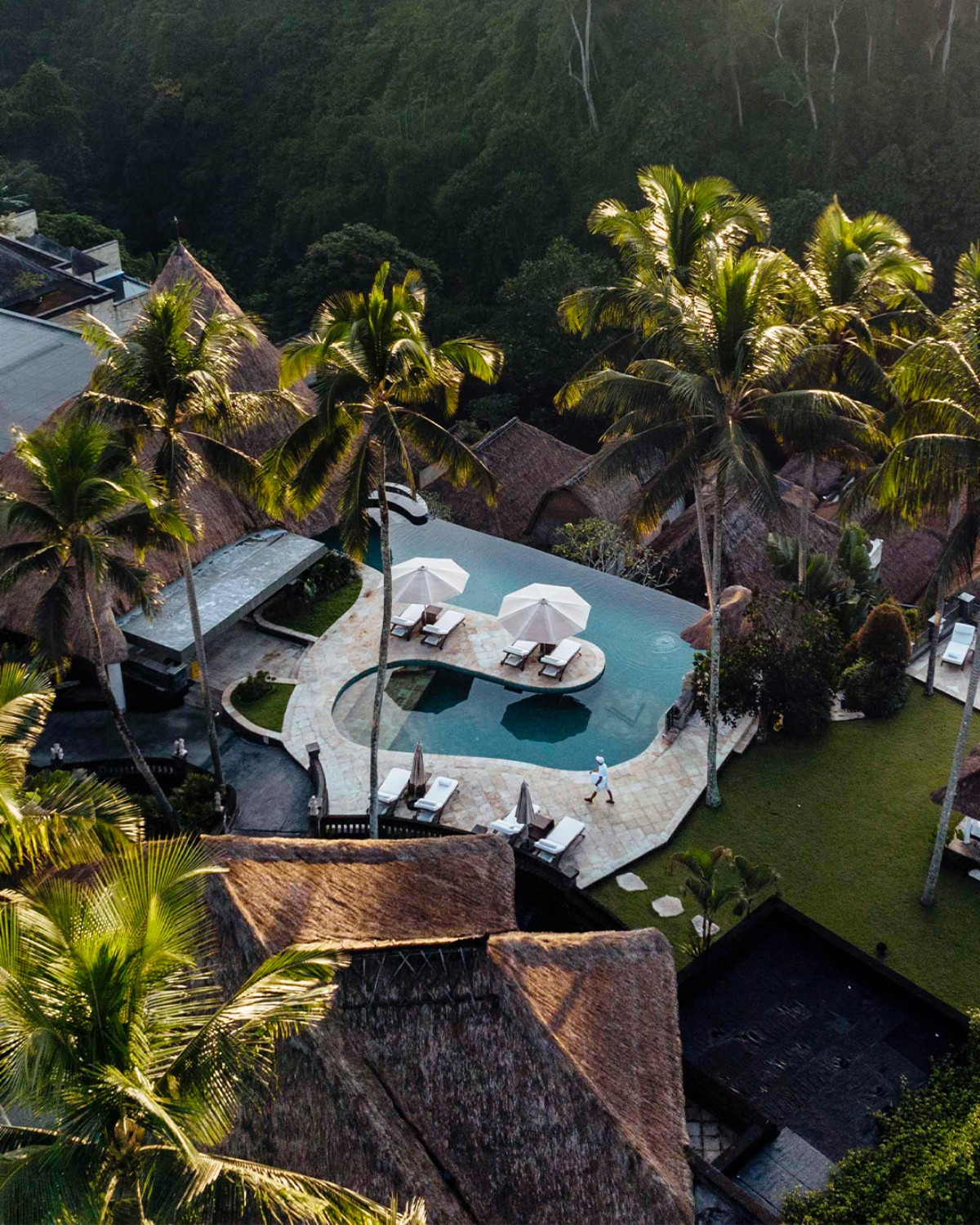 An elevated view of a luxurious resort with thatched roofs, a pristine pool, and the jungle backdrop.